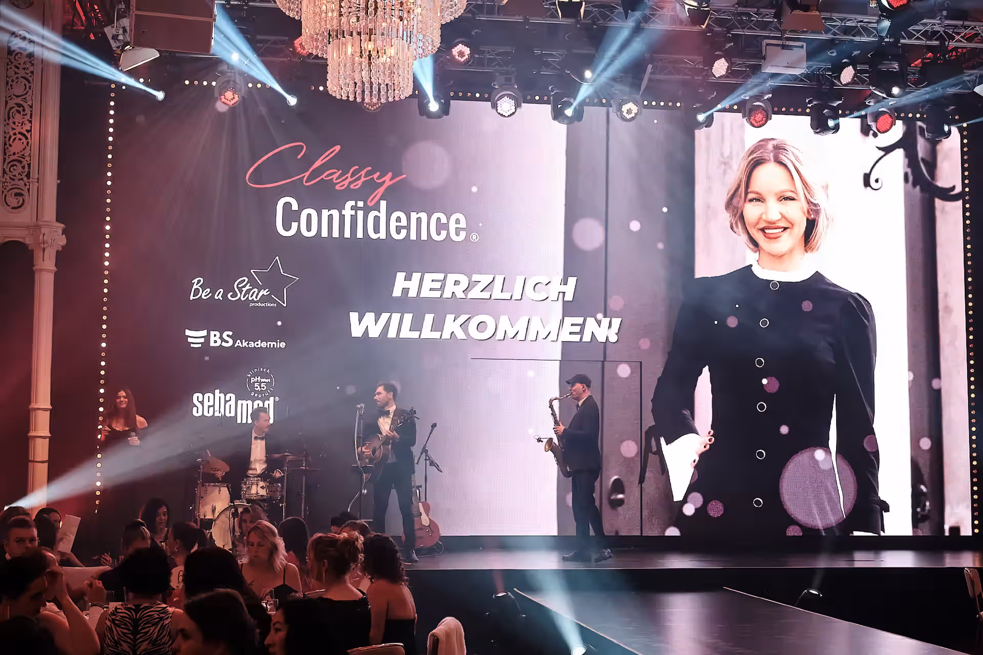 Frau in schwarzem Blazer steht vor Publikum und präsentiert mit Confidence-Schriftzug im Hintergrund