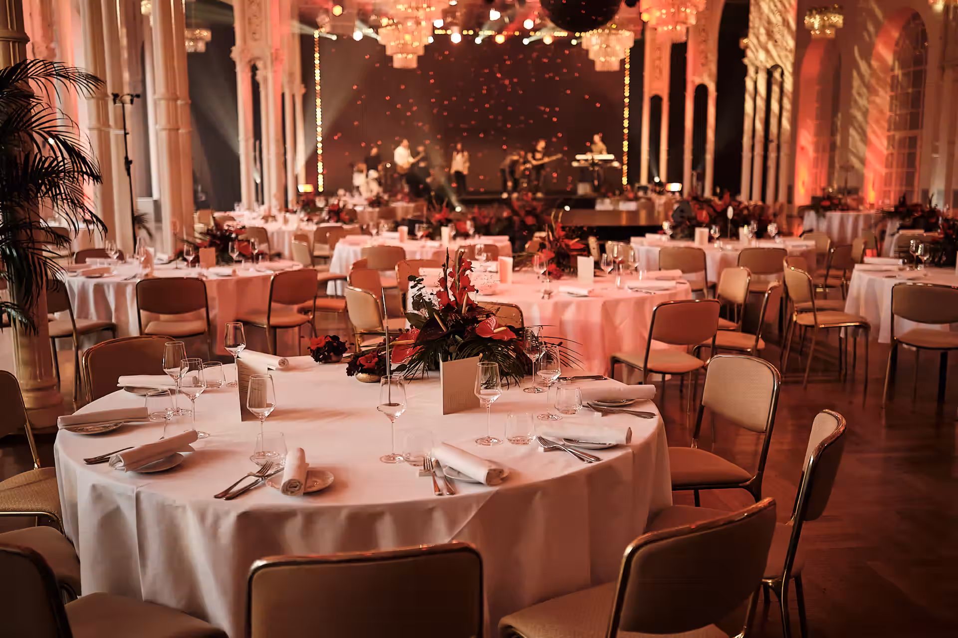 Eleganter Bankettsaal mit festlich gedeckten runden Tischen, weißen Tischdecken und roten Blumenarrangements, im Hintergrund Bühne mit Musikern.
