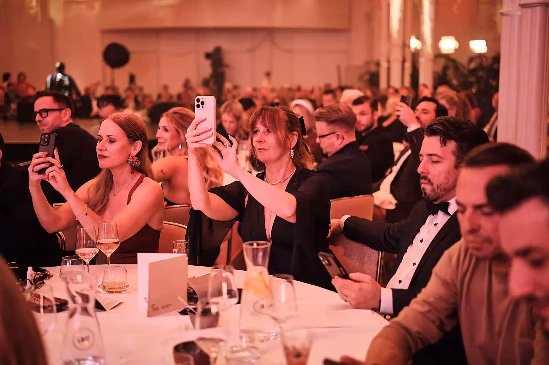 Menschen bei formeller Veranstaltung sitzen an Tisch, einige machen Fotos mit Smartphones.