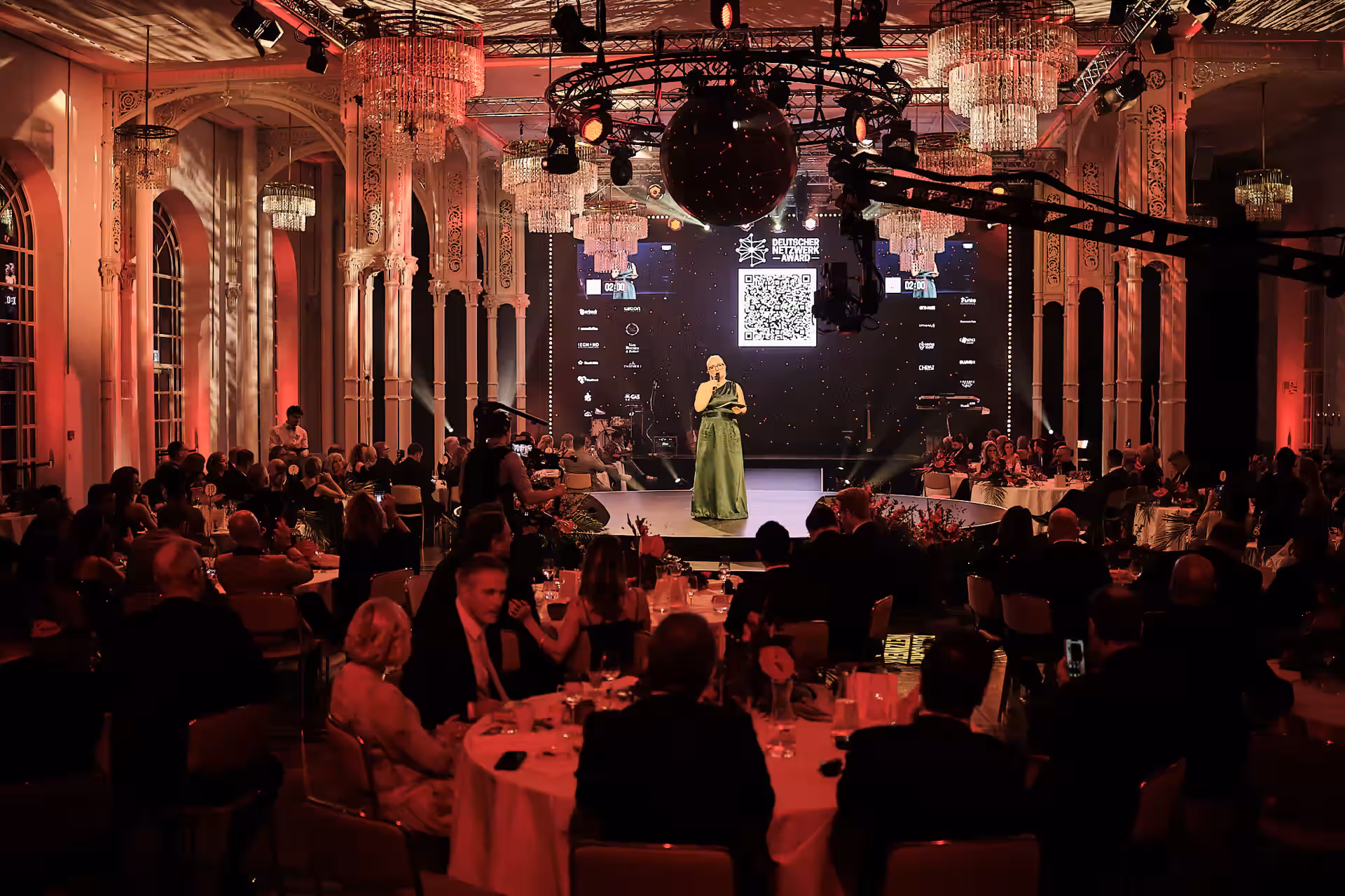 Frau im langen grünen Kleid spricht auf Bühne bei elegantem Galaevent mit Publikum an gedeckten Tischen in prunkvollem Saal.