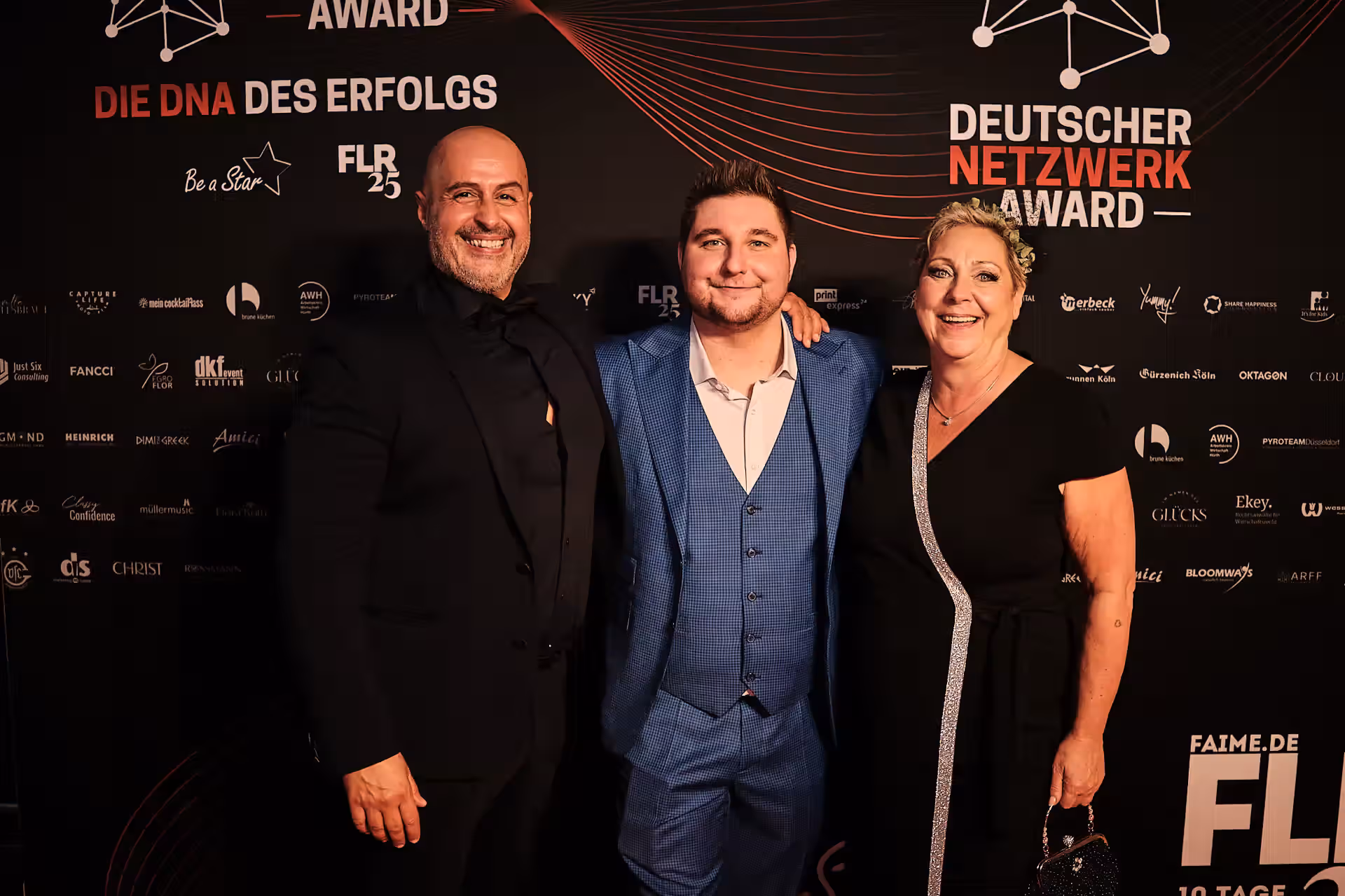 Drei lächelnde Personen posieren vor einer schwarzen Wand mit dem Schriftzug Deutscher Netzwerk Award und Logos verschiedener Sponsoren.