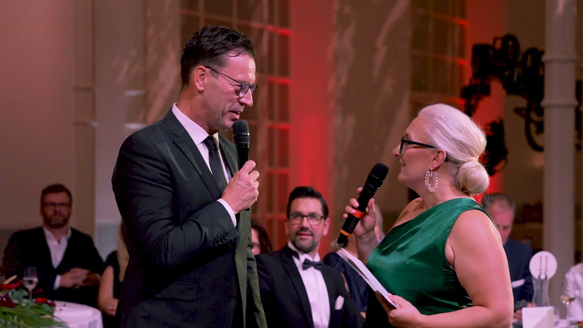 Mann im Anzug und Frau im grünen Kleid bei einer festlichen Veranstaltung mit Gästen im Hintergrund
