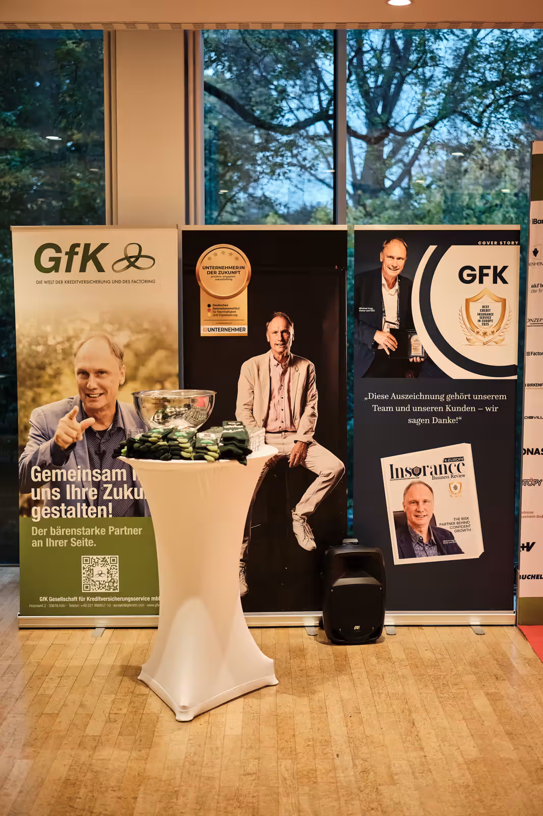 Messestand mit Bannern der GfK mit Bildern eines Mannes, die GfK-Logo, Auszeichnungen und ein Stehtisch mit Giveaways.
