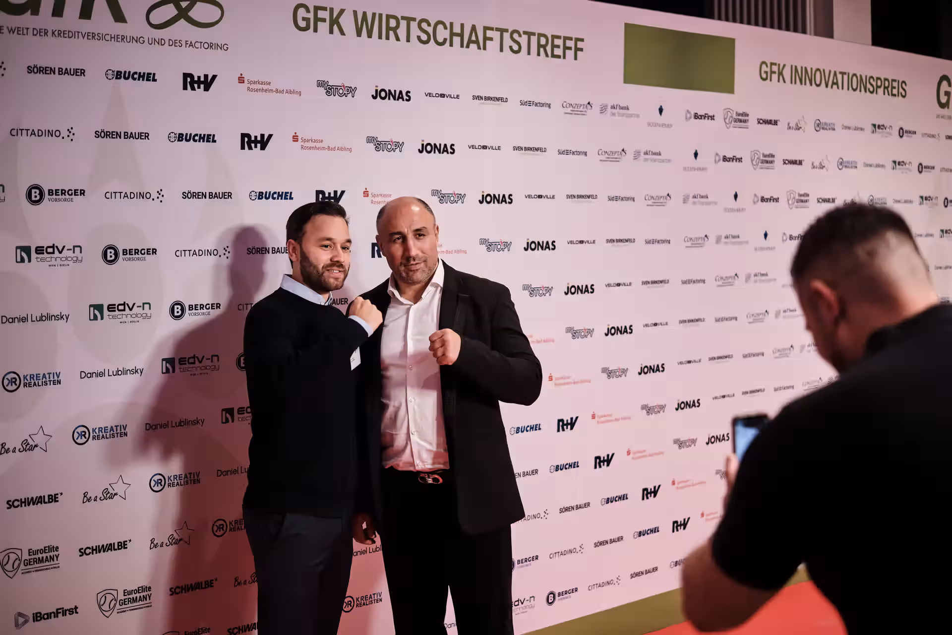 Zwei Männer posieren mit erhobenen Fäusten vor einer Sponsorenwand beim GFK Wirtschaftstreff, während eine dritte Person ein Foto von ihnen macht.