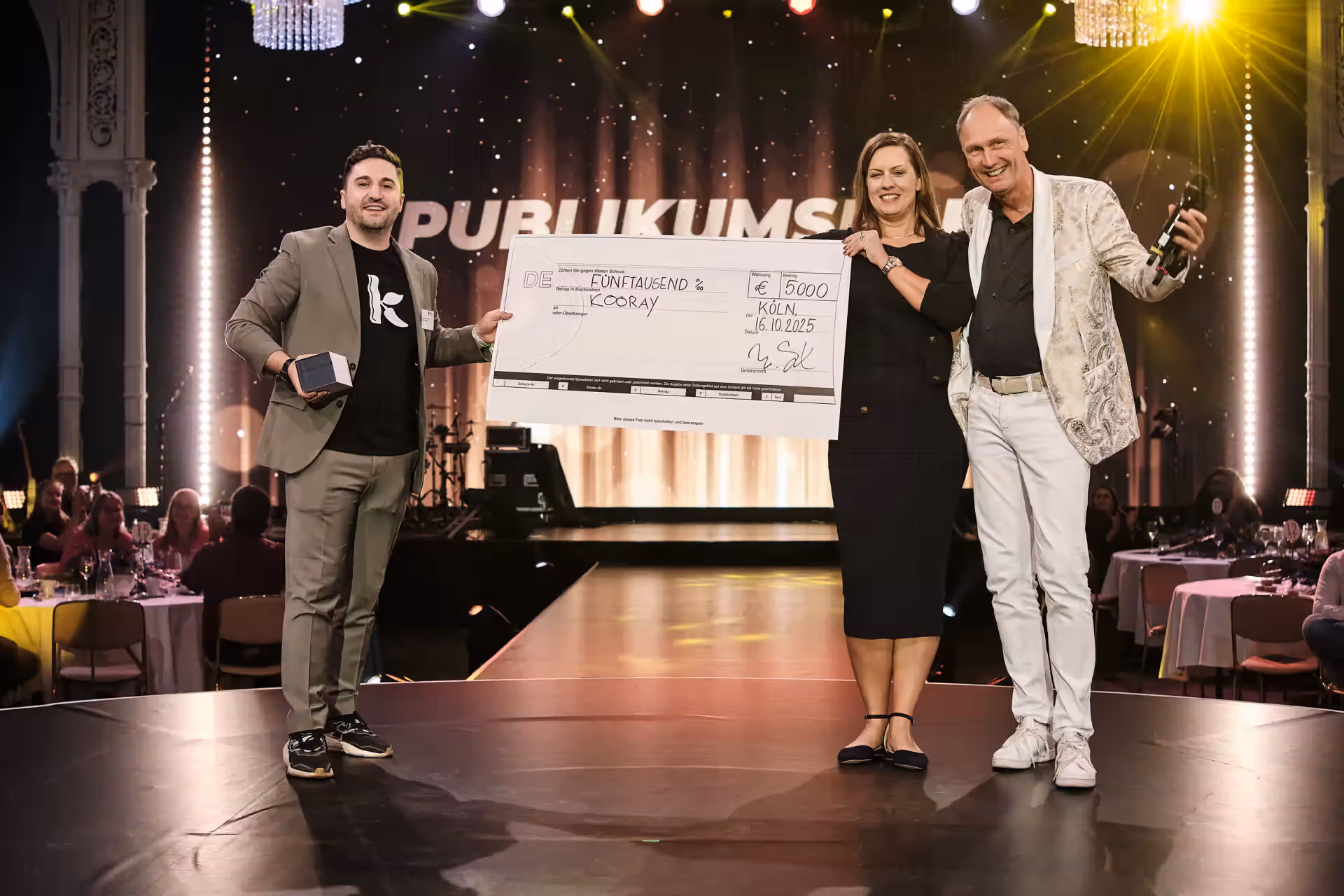 Drei Personen auf einer Bühne halten einen großen Scheck über 5000 Euro, umgeben von einem angesagten Veranstaltungshintergrund mit Publikum.