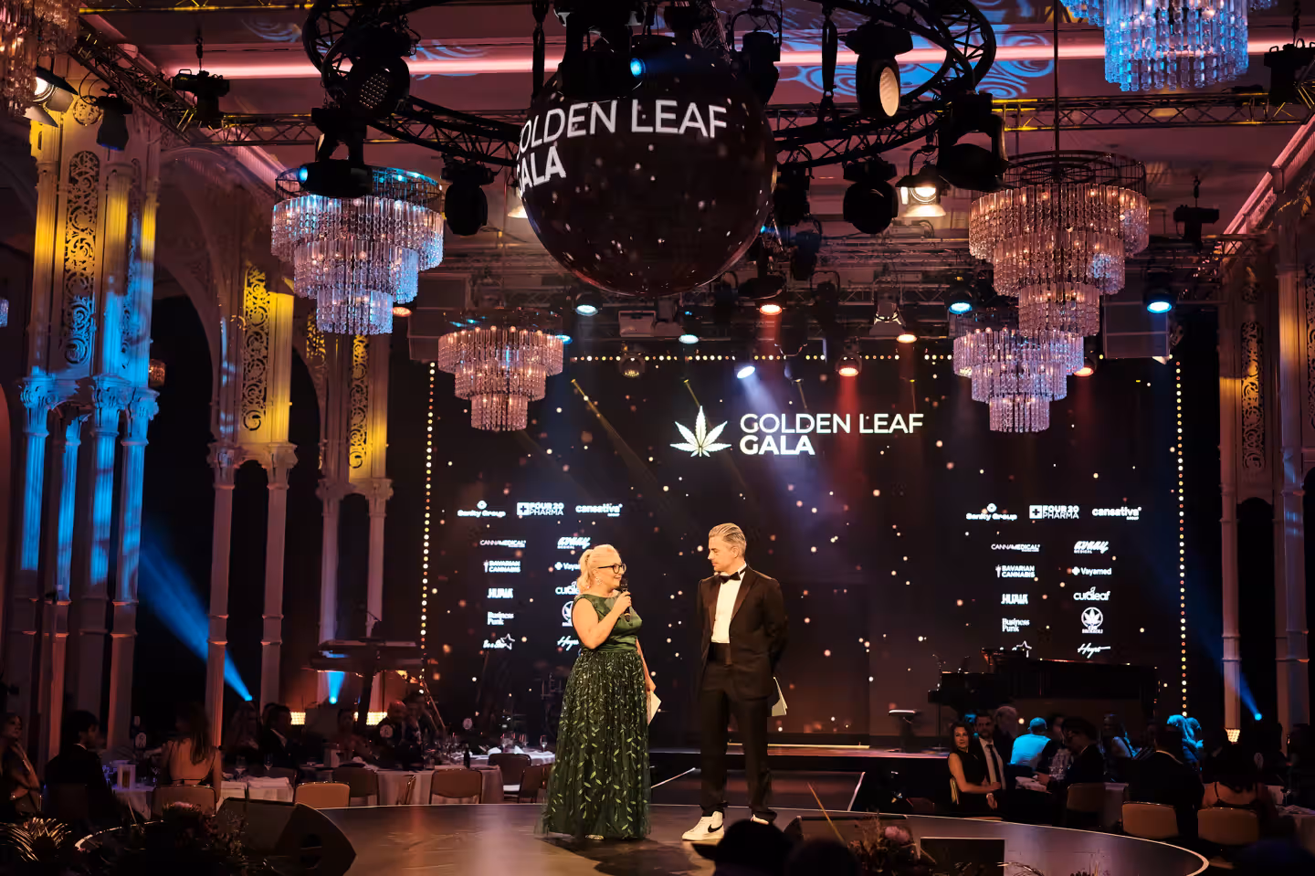 Bühne mit Moderatorenpaar bei der Golden Leaf Gala in einem prunkvollen Saal mit Lichterketten und Logos an den Wänden