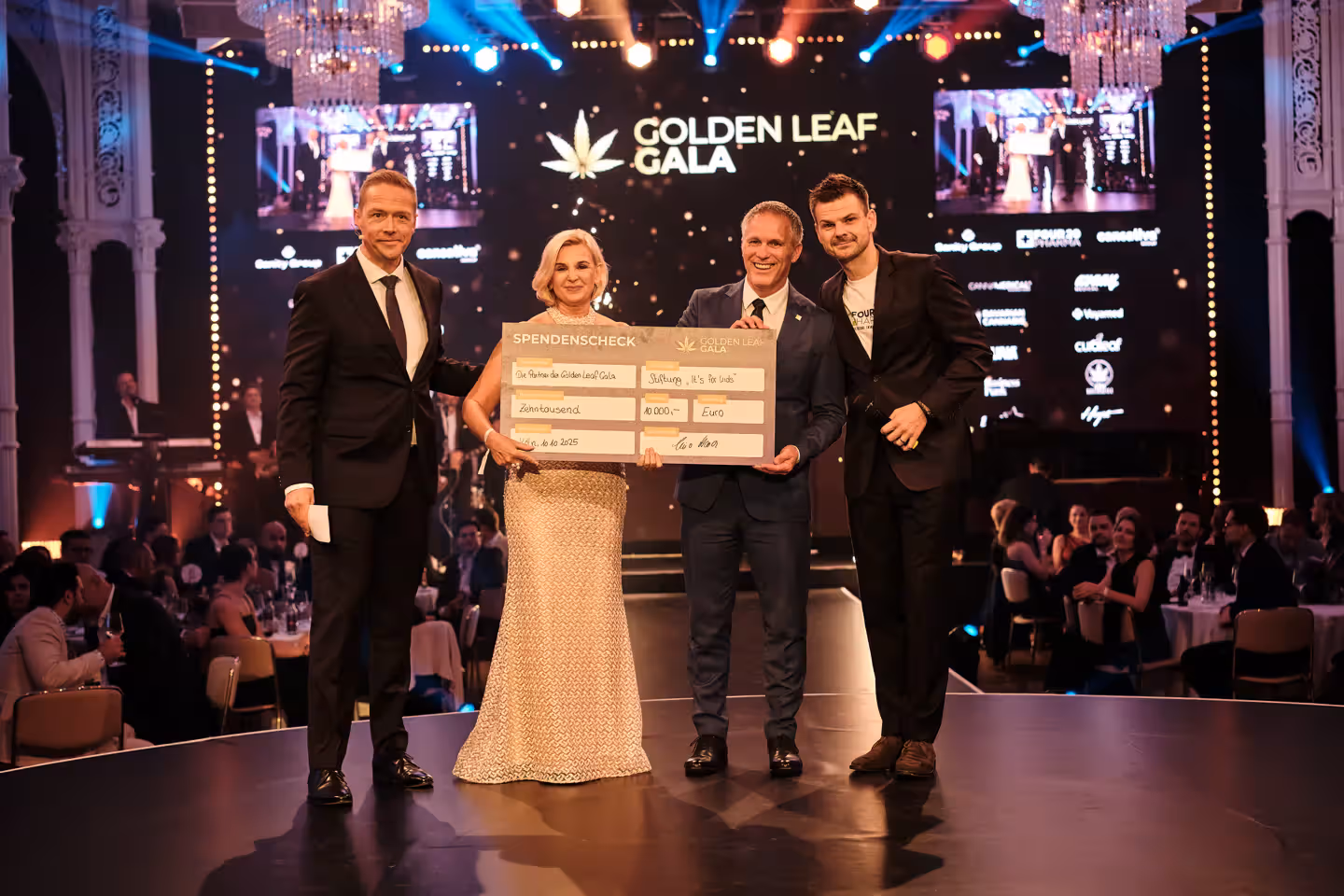 Drei Personen halten einen Spendenscheck bei der Golden Leaf Gala, umgeben von Gästen und Lichtern.