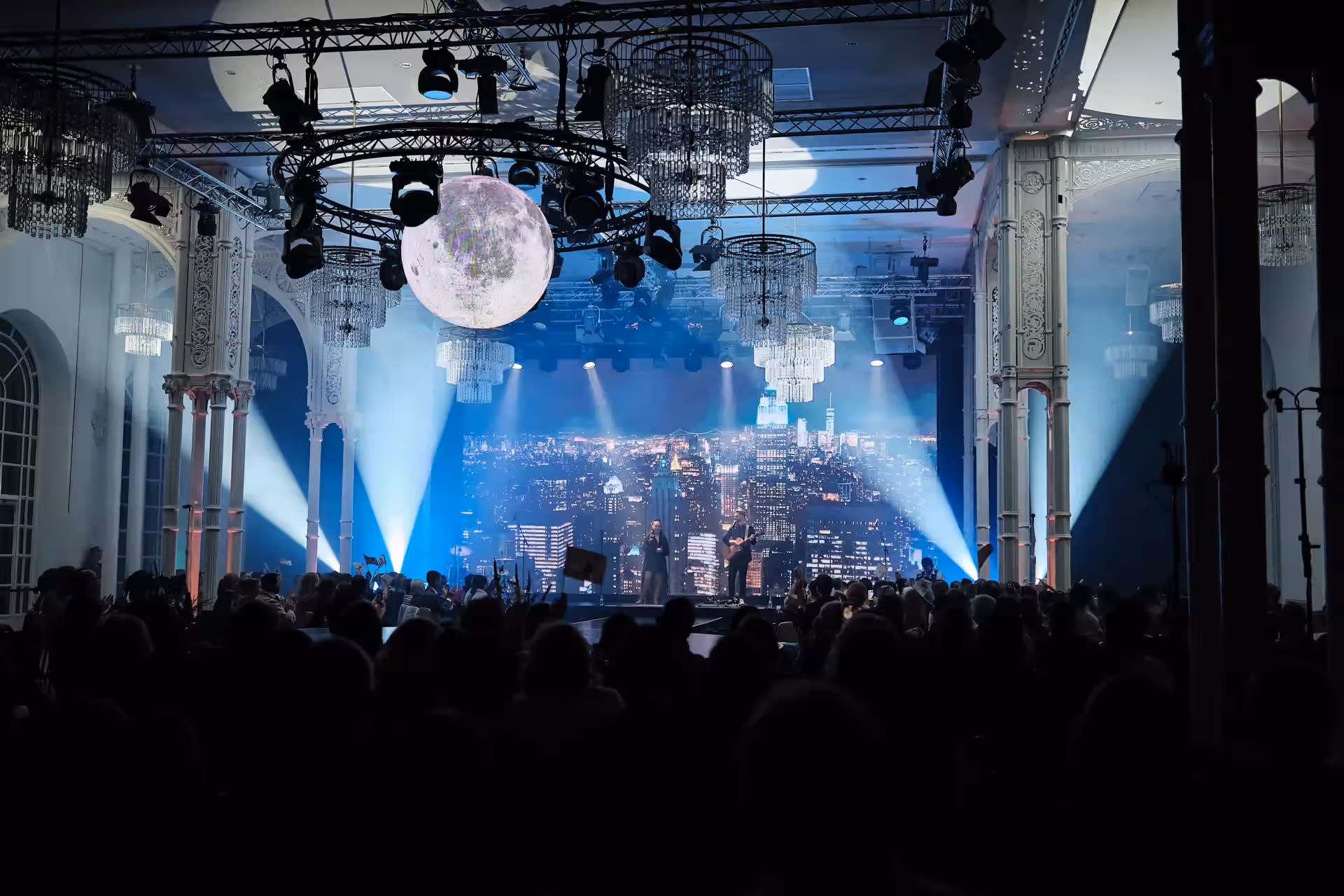 Große Indoor-Veranstaltung mit beleuchteter Bühne und Publikum vor moderner Stadtkulisse